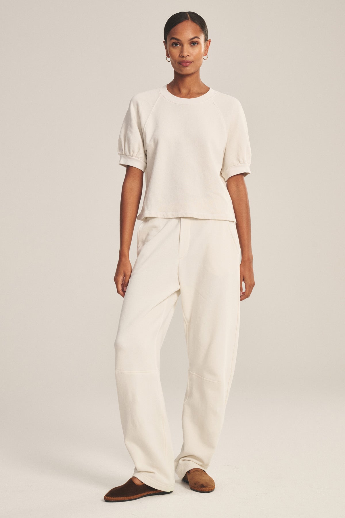 A woman stands against a plain background wearing a short-sleeve cream top and loose cream ELISE SWEATPANT by Velvet by Graham & Spencer, paired with brown slip-on shoes.-41038249623745