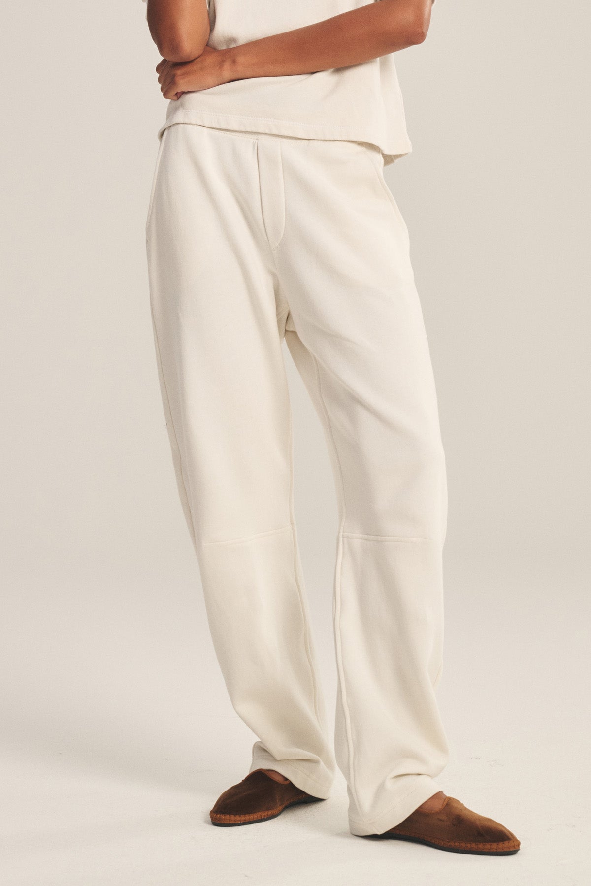 A person stands on a light background, arms crossed, wearing the ELISE SWEATPANT by Velvet by Graham & Spencer with a matching loose off-white lounge top and brown slippers.-41038249558209