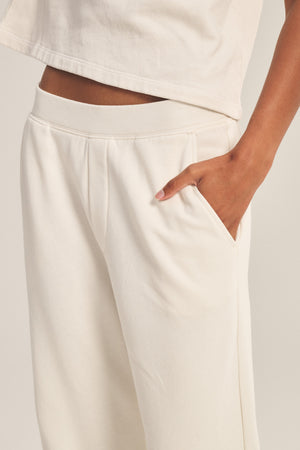 A person wearing Velvet by Graham & Spencer's ELISE SWEATPANT in a cream shade stands with one hand in their pocket, paired with a matching top.