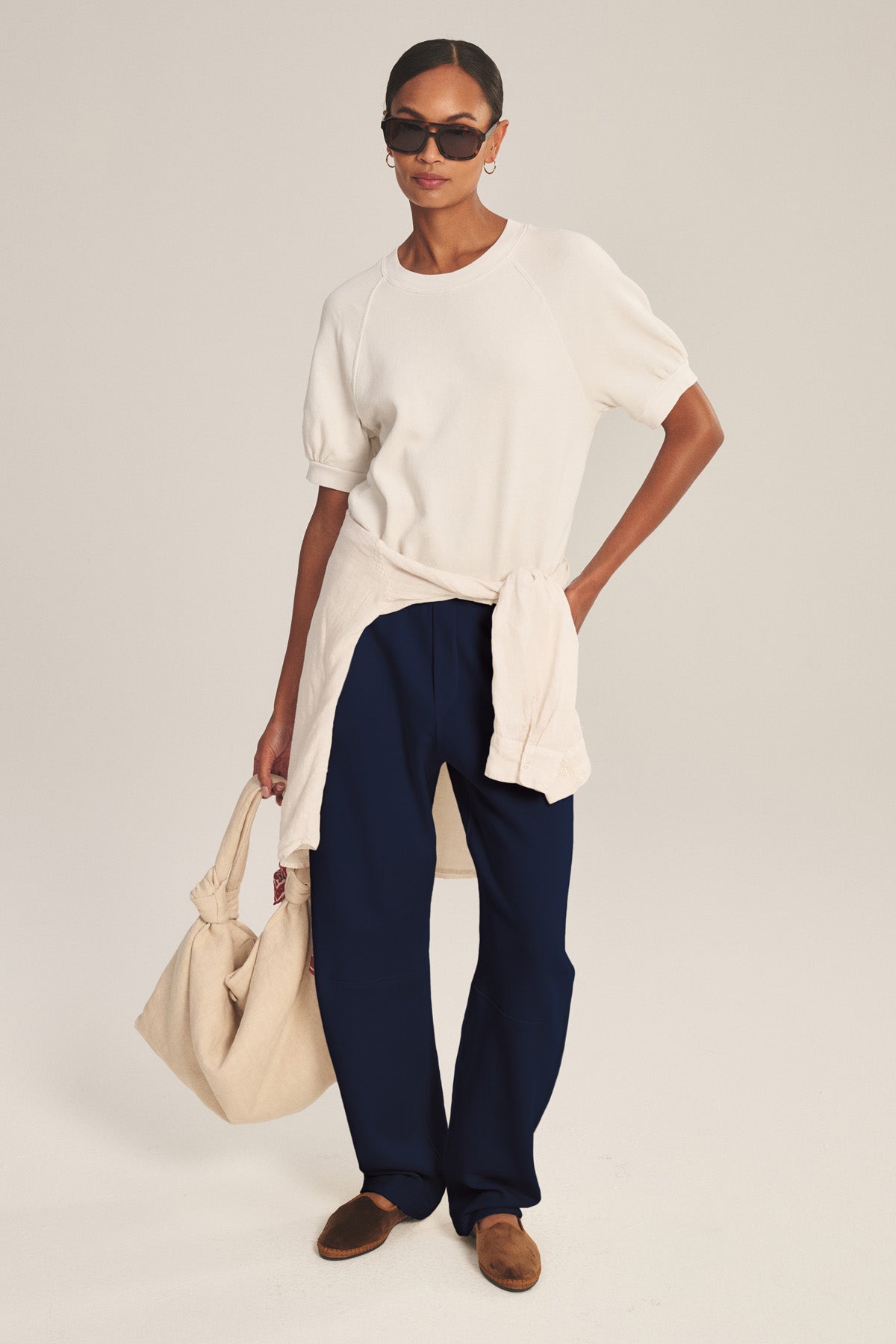 Wearing Velvet by Graham & Spencer's ELISE SWEATPANT in navy blue, a white short-sleeve top, brown shoes, sunglasses, hoop earrings, a cream sweater tied at the waist, and holding a beige tote bag.