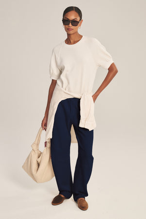 Wearing Velvet by Graham & Spencer's ELISE SWEATPANT in navy blue, a white short-sleeve top, brown shoes, sunglasses, hoop earrings, a cream sweater tied at the waist, and holding a beige tote bag.