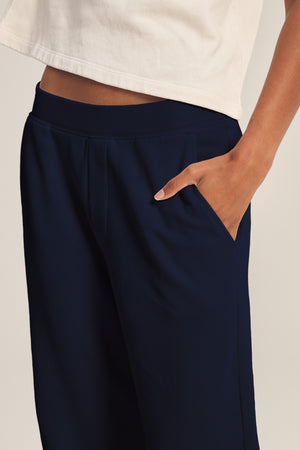 A person wearing a white top and Velvet by Graham & Spencer's ELISE SWEATPANT in navy blue cotton fleece stands with one hand in their pocket.