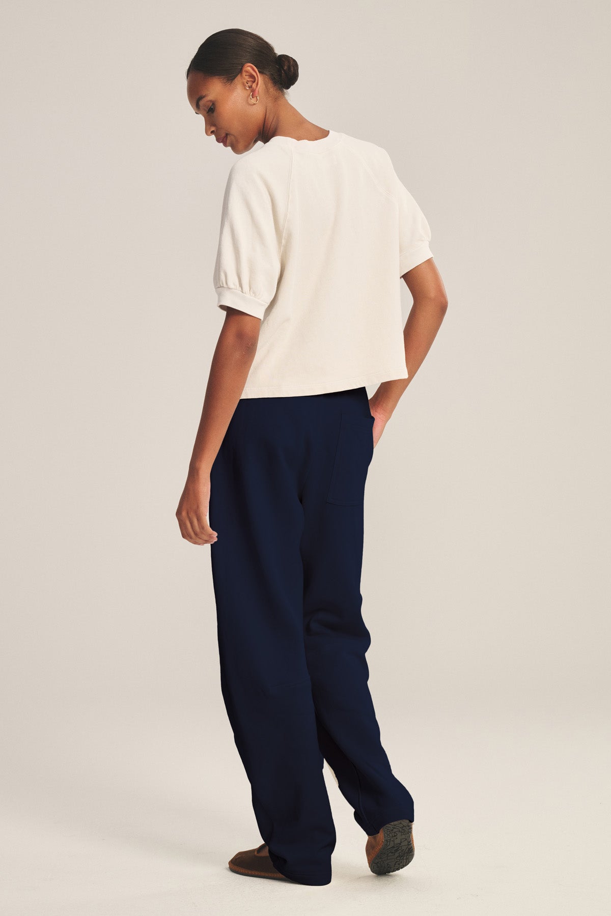A person stands slightly turned away, hair tied back, wearing Velvet by Graham & Spencer's ELISE SWEATPANT in dark blue with a relaxed barrel-leg silhouette and a white short-sleeve top, set against a plain light background.