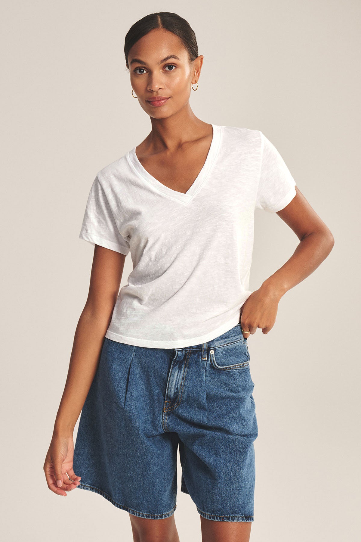 A woman wears the ELISHA TEE by Velvet by Graham & Spencer—a classic white V-neck in soft cotton slub—paired with blue denim shorts, standing against a plain background with one hand in her pocket.-41629729226945