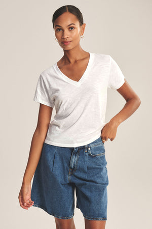 A woman wears the ELISHA TEE by Velvet by Graham & Spencer—a classic white V-neck in soft cotton slub—paired with blue denim shorts, standing against a plain background with one hand in her pocket.