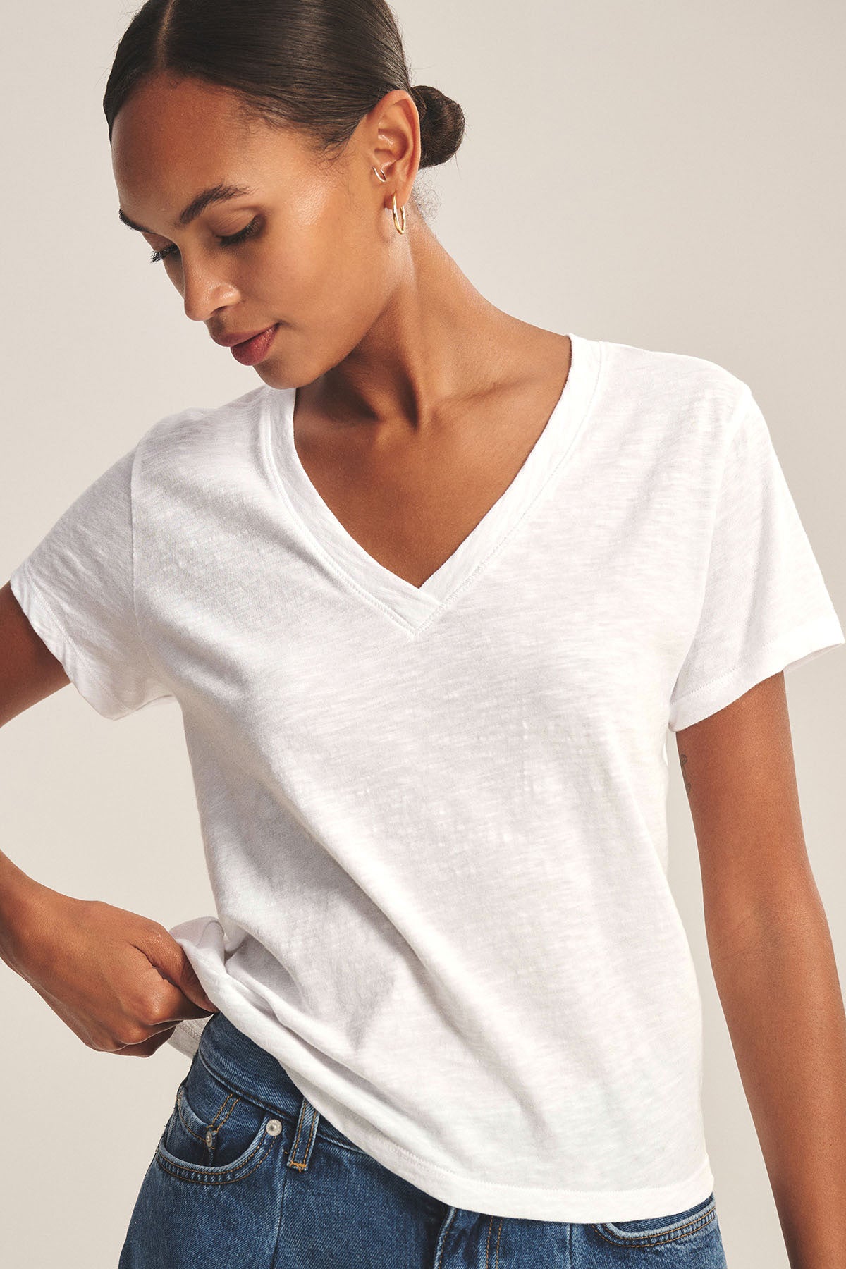 A woman wears the ELISHA TEE by Velvet by Graham & Spencer—a classic cotton slub V-neck tee—paired with blue jeans, looking down and holding the side of her shirt, showcasing this timeless wardrobe staple.