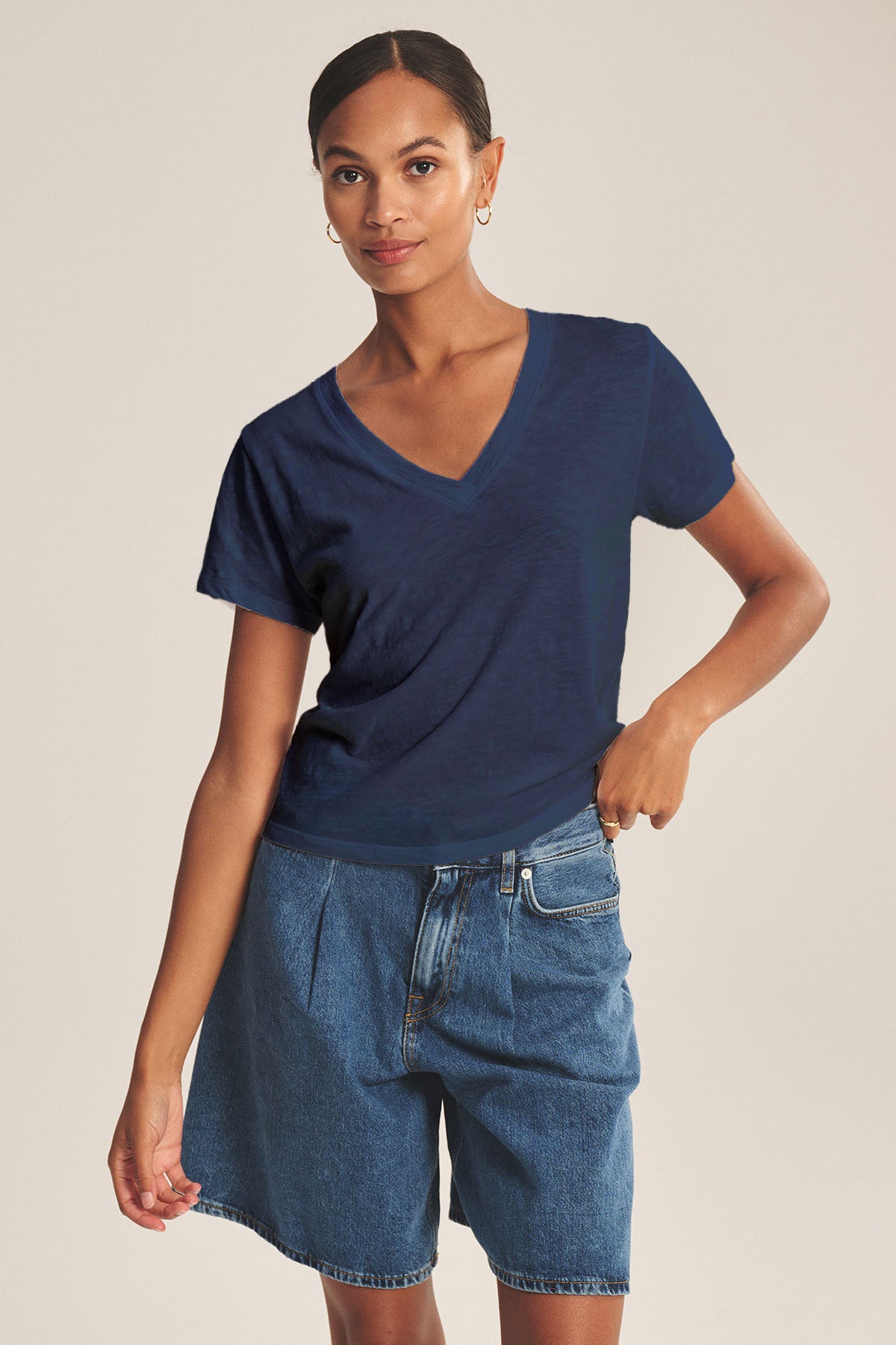 A woman stands against a plain background, wearing the ELISHA TEE by Velvet by Graham & Spencer—a navy cotton slub V-neck paired with high-waisted blue denim shorts for a timeless look.