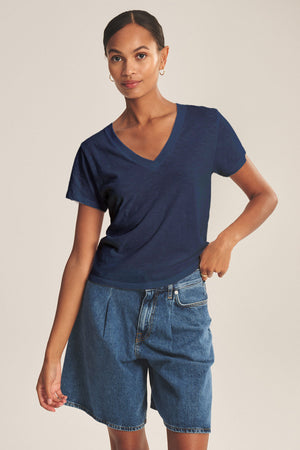 A woman stands against a plain background, wearing the ELISHA TEE by Velvet by Graham & Spencer—a navy cotton slub V-neck paired with high-waisted blue denim shorts for a timeless look.
