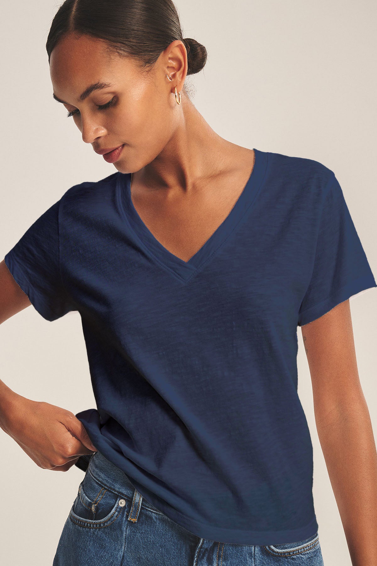 A woman models the ELISHA TEE by Velvet by Graham & Spencer, a navy blue cotton slub V-neck, paired with blue jeans as she stands against a neutral background, looking down with one hand in her pocket.