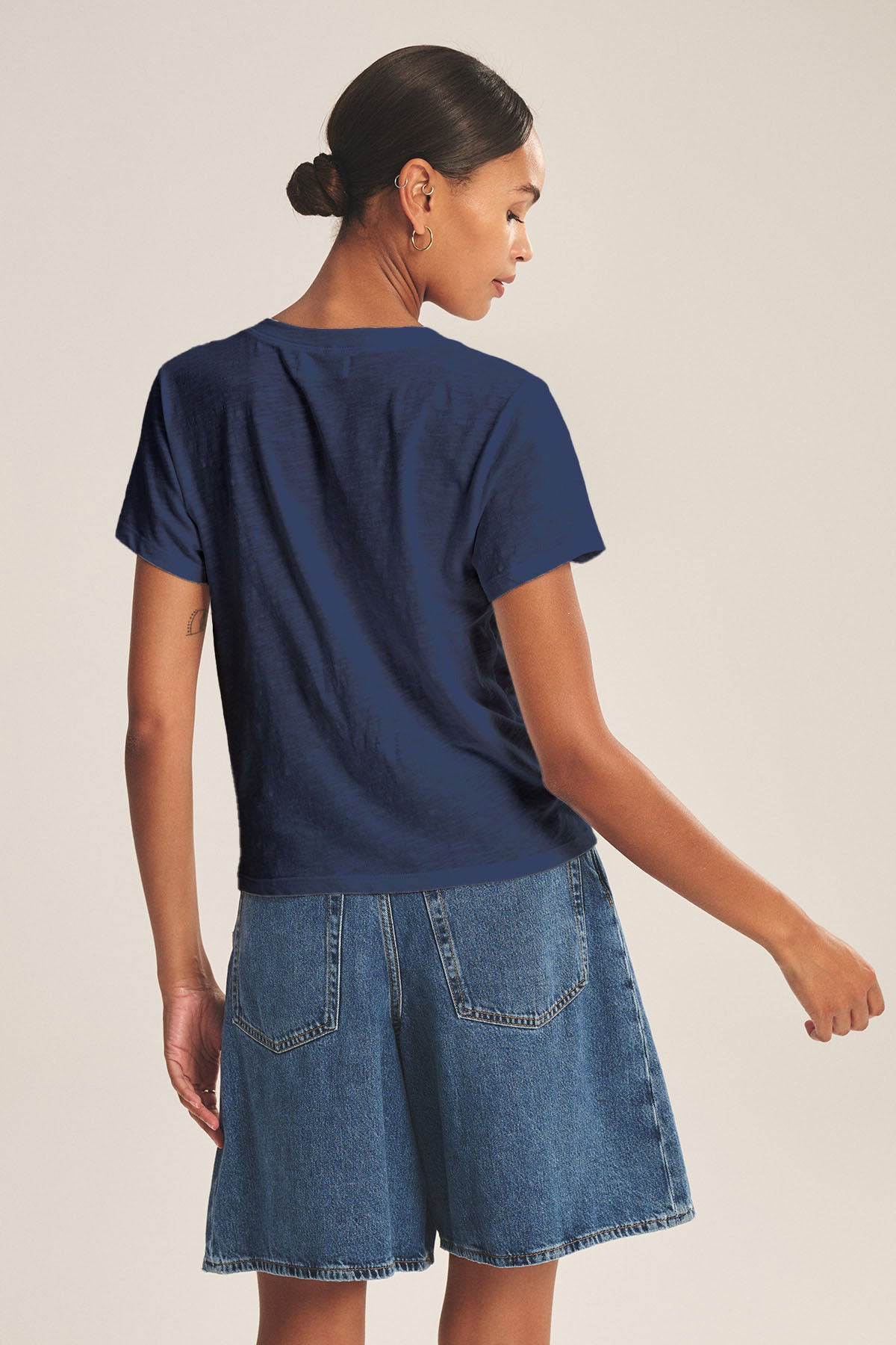 A woman faces away, wearing the ELISHA TEE by Velvet by Graham & Spencer—a navy cotton slub V-neck—styled with high-waisted denim shorts and her hair in a low bun.