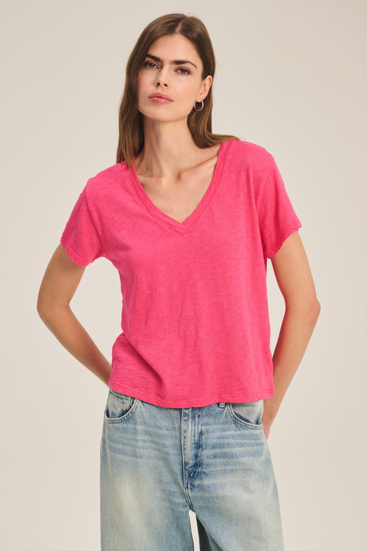 A woman stands against a plain light background wearing the Velvet by Graham & Spencer ELISHA TEE in bright pink cotton slub and light blue jeans.