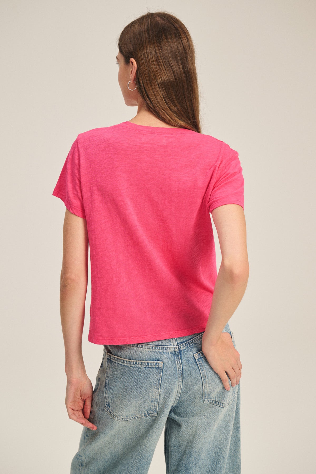 A person stands with their back to the camera, wearing the ELISHA TEE by Velvet by Graham & Spencer in bright pink cotton slub and light blue jeans—a perfect wardrobe staple.-41178042007745
