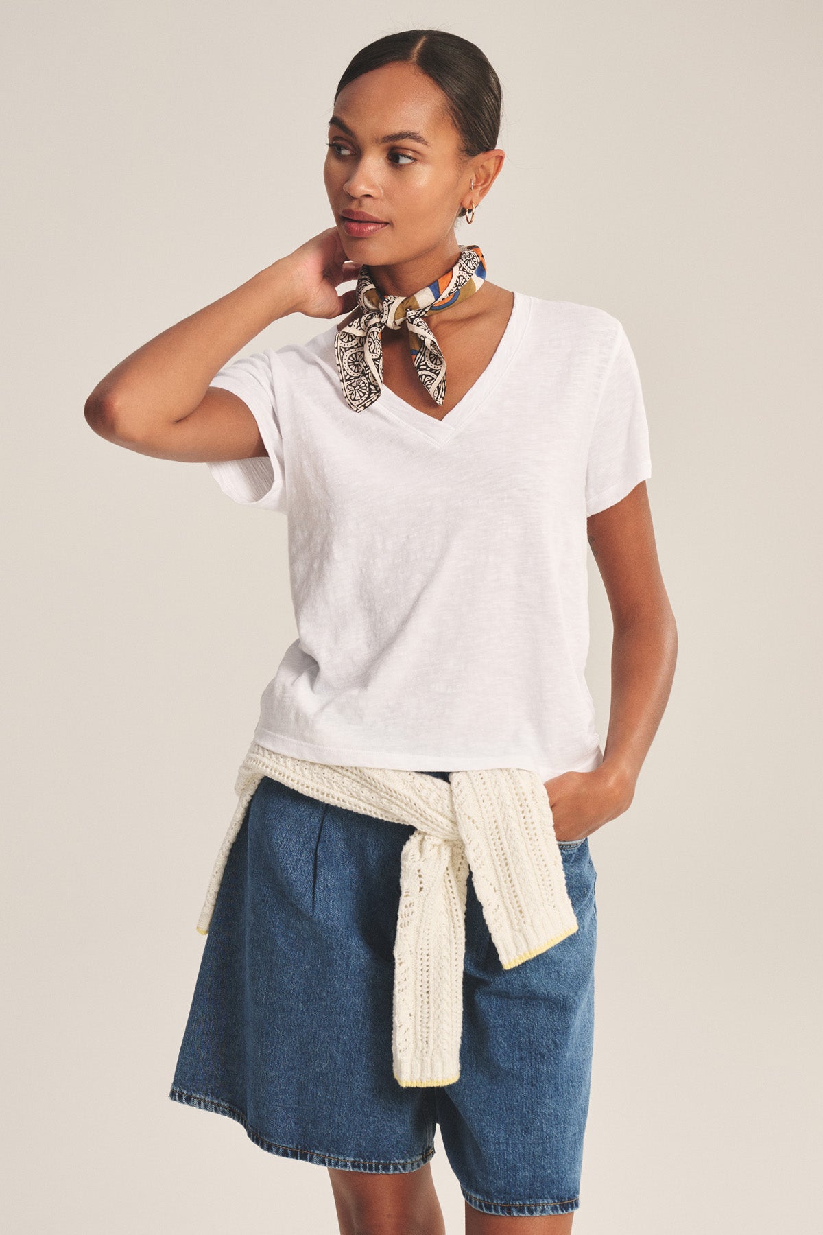 A woman stands with one hand behind her neck, wearing the ELISHA TEE by Velvet by Graham & Spencer, denim shorts, a cream sweater tied at the waist, and a patterned scarf around her neck.