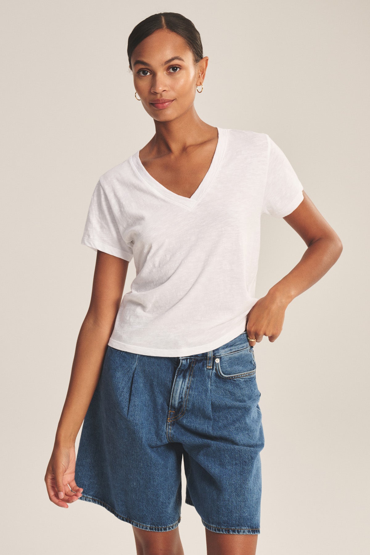 A woman stands against a plain background wearing the ELISHA TEE by Velvet by Graham & Spencer—a white V-neck tee styled with blue denim shorts and one hand in her pocket, making it a true wardrobe staple.