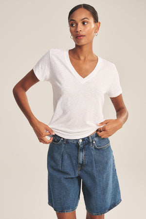 A person wears the ELISHA TEE by Velvet by Graham & Spencer, a classic V-neck crafted from soft cotton slub, paired with knee-length blue denim shorts, standing hands on waist against a neutral background.