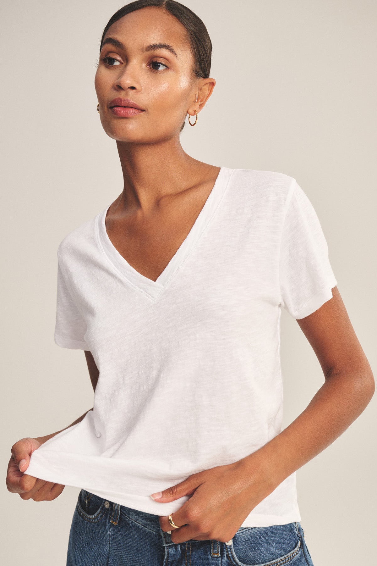 A person wearing the ELISHA TEE by Velvet by Graham & Spencer in white stands against a plain background, paired with blue jeans and holding the shirt's hem with one hand.-41044728512705