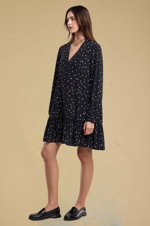 A woman stands in profile wearing the ELSA SILK DRESS by Velvet by Graham & Spencer—a loose, long-sleeved black silk dress with white polka dots, featuring a V-neckline and a ruffled, tiered skirt—paired with black loafers.