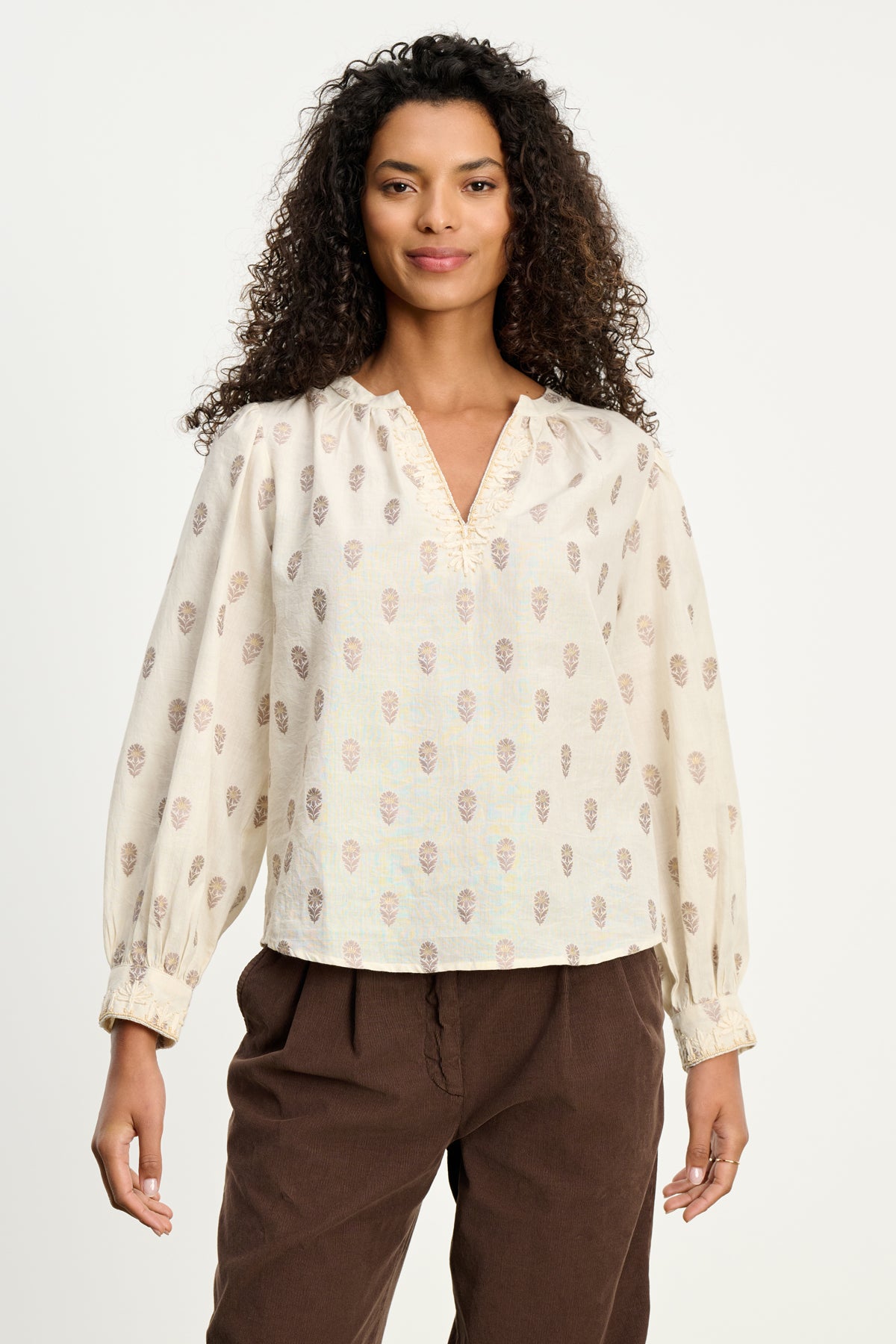   A woman with curly hair wears the ANA TOP by Velvet by Graham & Spencer, a cream embroidered cotton blouse with a subtle pattern, paired with brown pants against a plain white background. 
