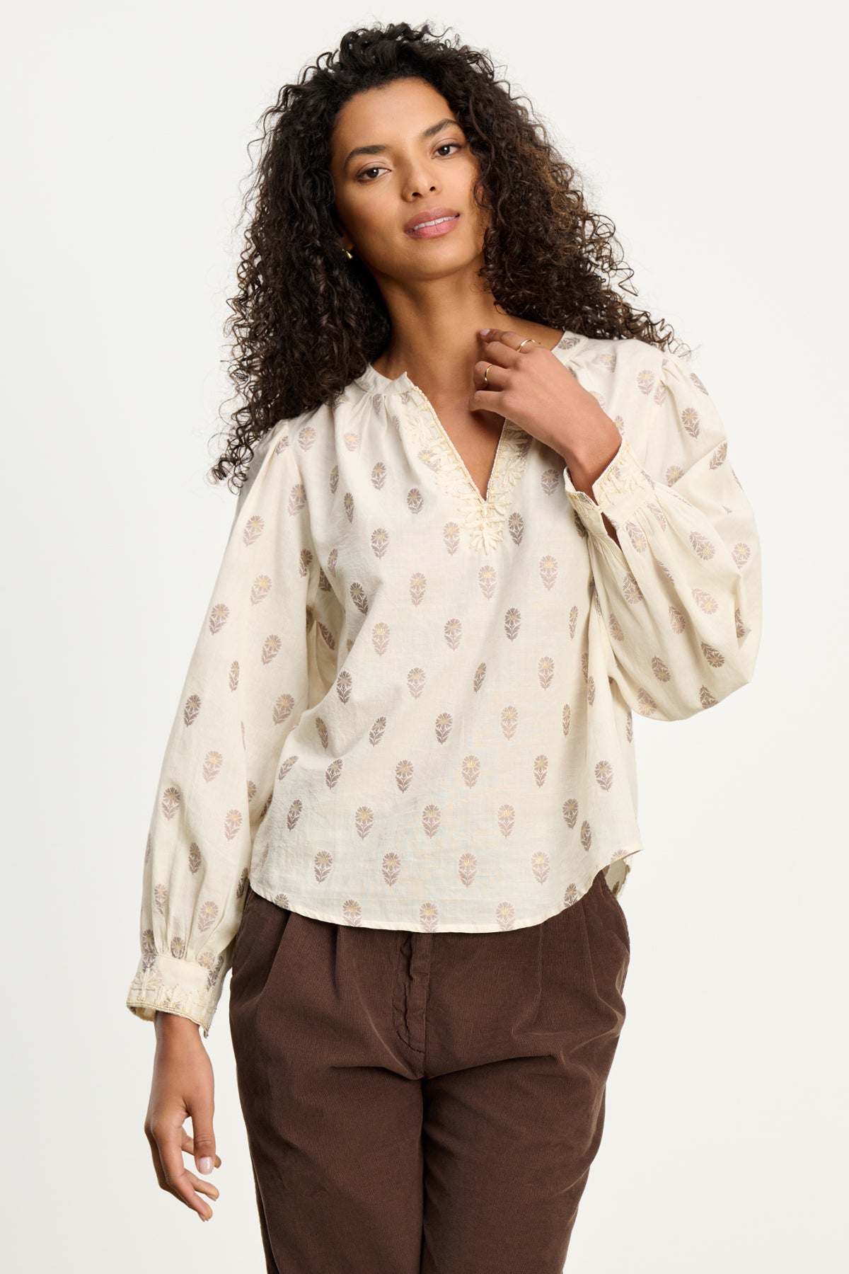   A woman with curly hair wears the ANA TOP by Velvet by Graham & Spencer, a cream embroidered cotton blouse with a subtle print, paired with brown pants against a plain light background. 