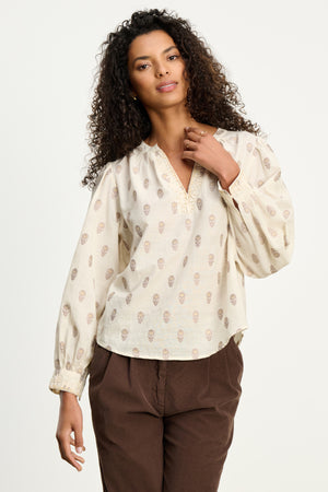A woman with curly hair wears the ANA TOP by Velvet by Graham & Spencer, a cream embroidered cotton blouse with a subtle print, paired with brown pants against a plain light background.