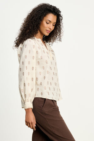 A woman with curly hair wears the ANA TOP by Velvet by Graham & Spencer, an embroidered cream cotton top with a subtle print, paired with brown pants and standing against a plain light background.