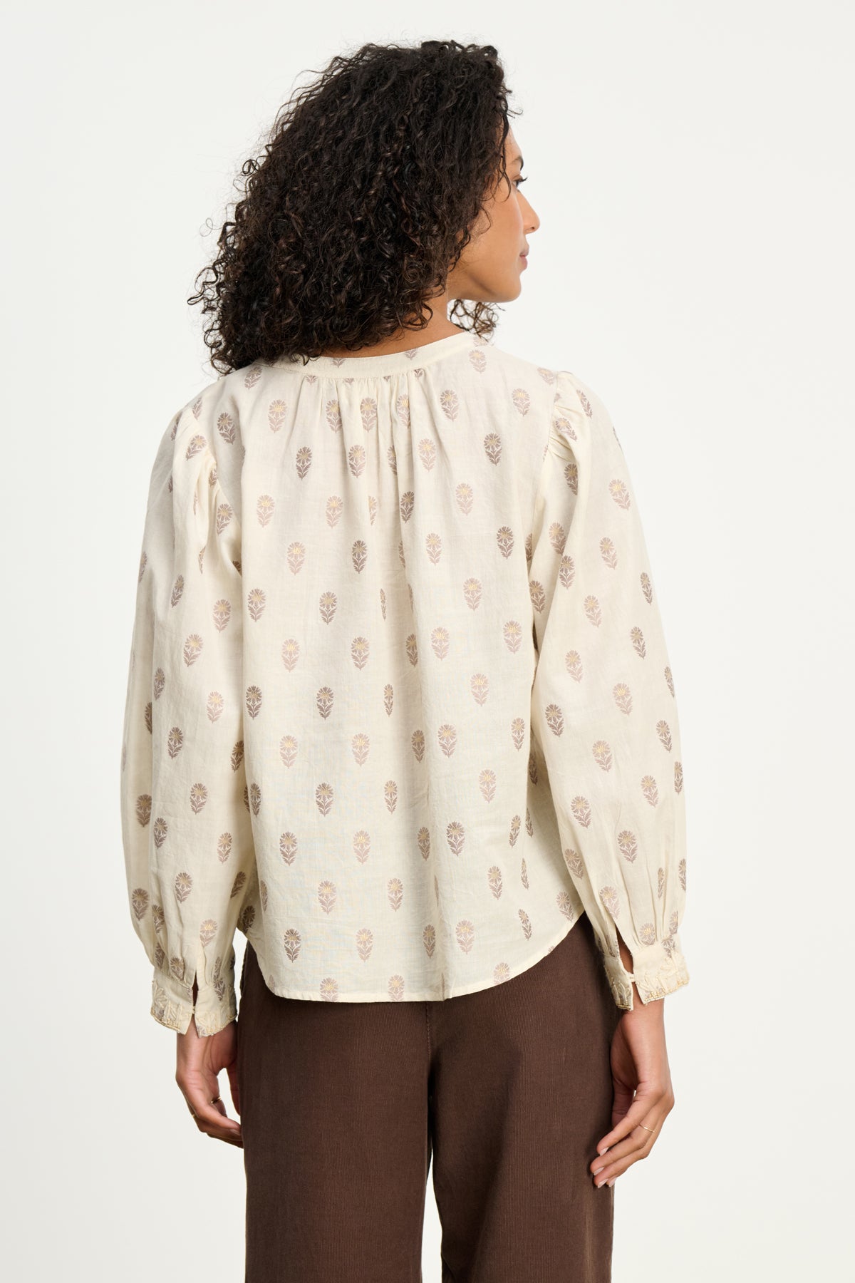A woman with curly hair is shown from the back wearing the ANA TOP by Velvet by Graham & Spencer—a light-colored, long-sleeve blouse with a subtle pattern—paired with brown pants against a plain background.-39617373896897