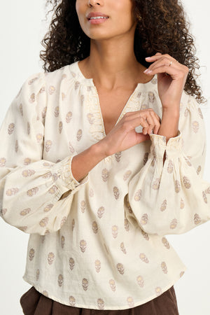 A woman stands against a plain background wearing the ANA TOP by Velvet by Graham & Spencer, featuring a cream hue, light brown leaf pattern, and split neckline.