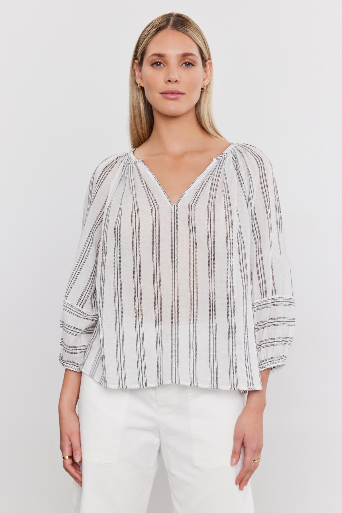   Woman wearing a Velvet by Graham & Spencer Gianna top, a striped v-neckline blouse with three-quarter sleeves, paired with white pants, standing against a white background. 