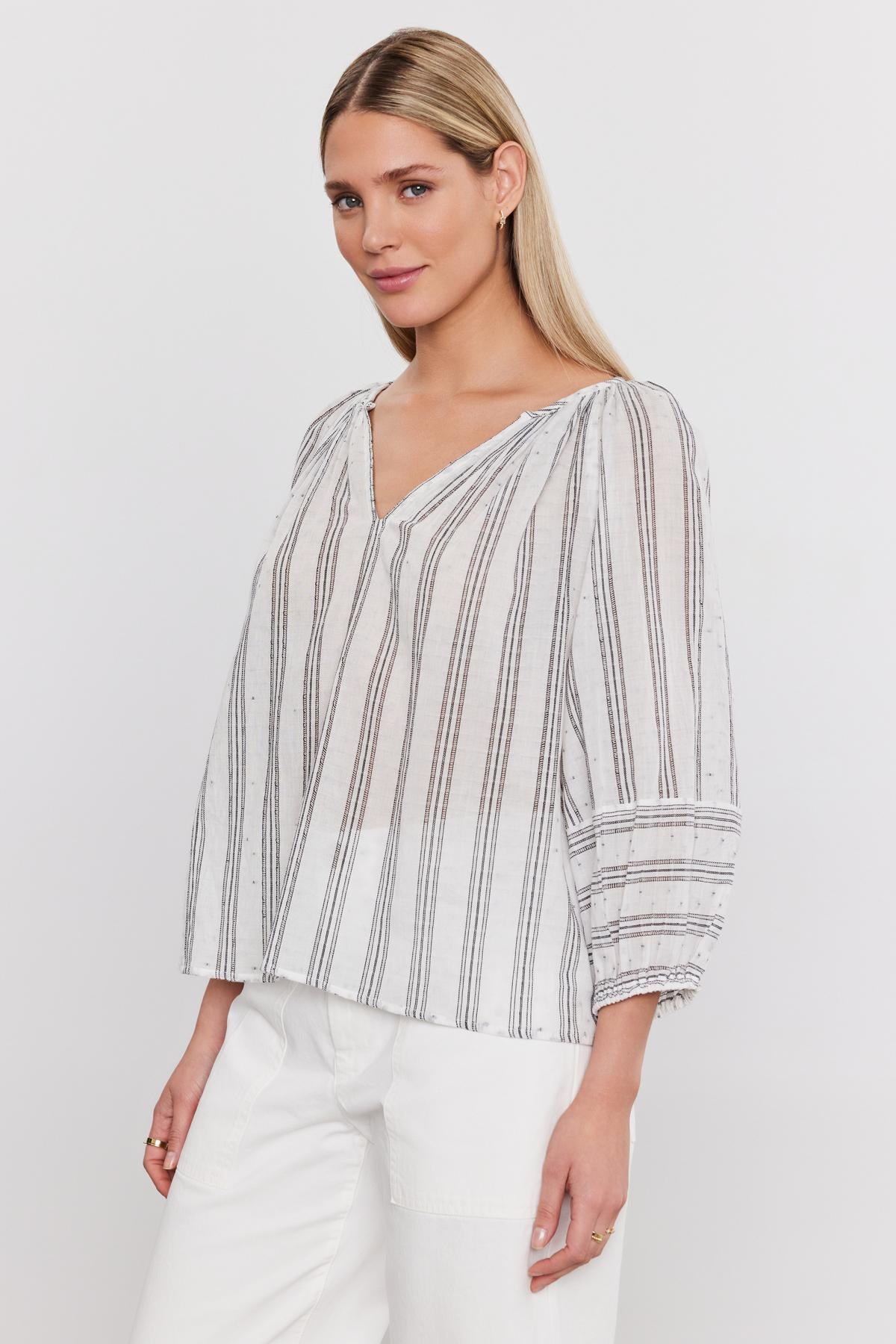  A woman models a white and grey striped cotton Gianna top with three-quarter sleeves and elastic cuffs, paired with white pants, against a plain background by Velvet by Graham & Spencer. 