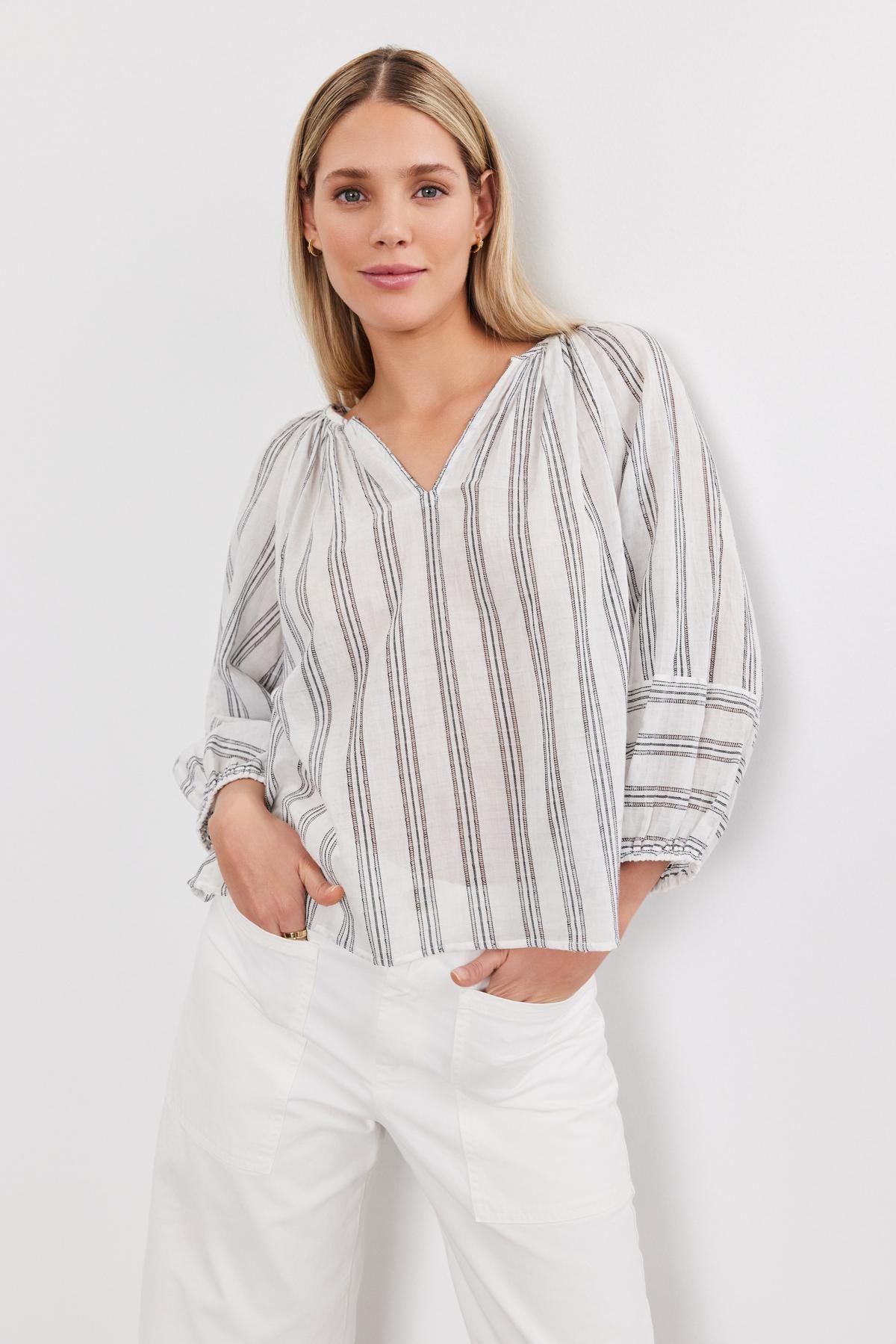   A woman stands confidently, wearing the Gianna top by Velvet by Graham & Spencer with vertical stripes and white pants, with her hand in her pocket. 