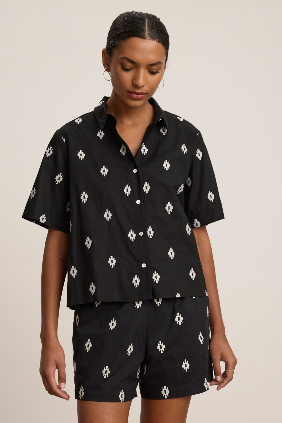 A person wearing the MACY TOP by Velvet by Graham & Spencer—a matching black short-sleeve button-up with a white geometric pattern—stands against a plain light background.-39488943882433