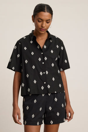 A person wearing the MACY TOP by Velvet by Graham & Spencer—a matching black short-sleeve button-up with a white geometric pattern—stands against a plain light background.