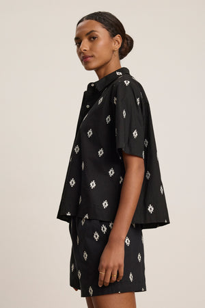 A woman stands sideways wearing the MACY TOP by Velvet by Graham & Spencer, a loose-fitting black short-sleeve shirt paired with matching shorts featuring a white geometric print.