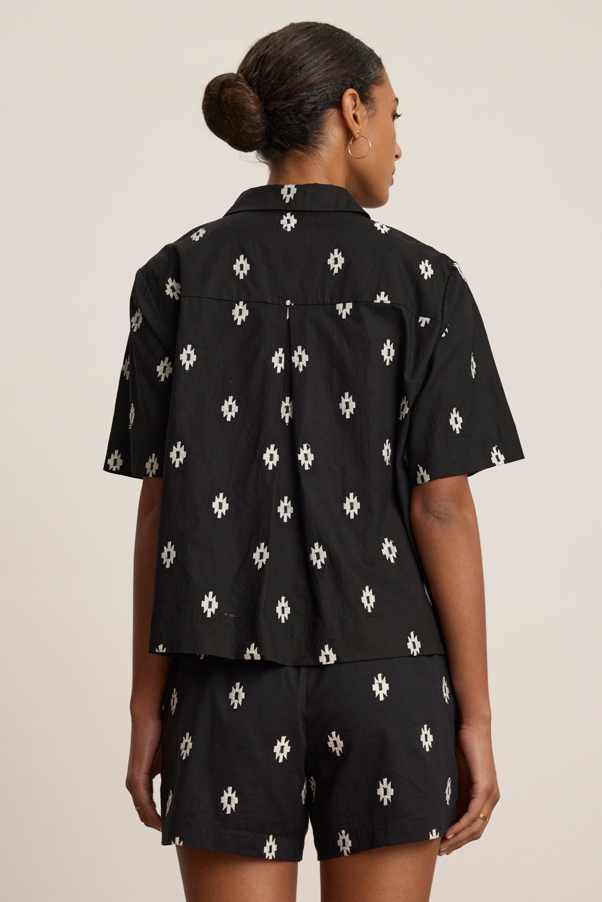 A woman faces away, wearing the Velvet by Graham & Spencer MACY TOP, a black short-sleeve shirt. Her hair is styled in a low bun.-39488943947969