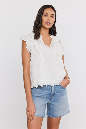 A woman in a white CHARLENE TOP with cotton embroidery and denim shorts, standing against a plain background. (Brand: Velvet by Graham & Spencer)