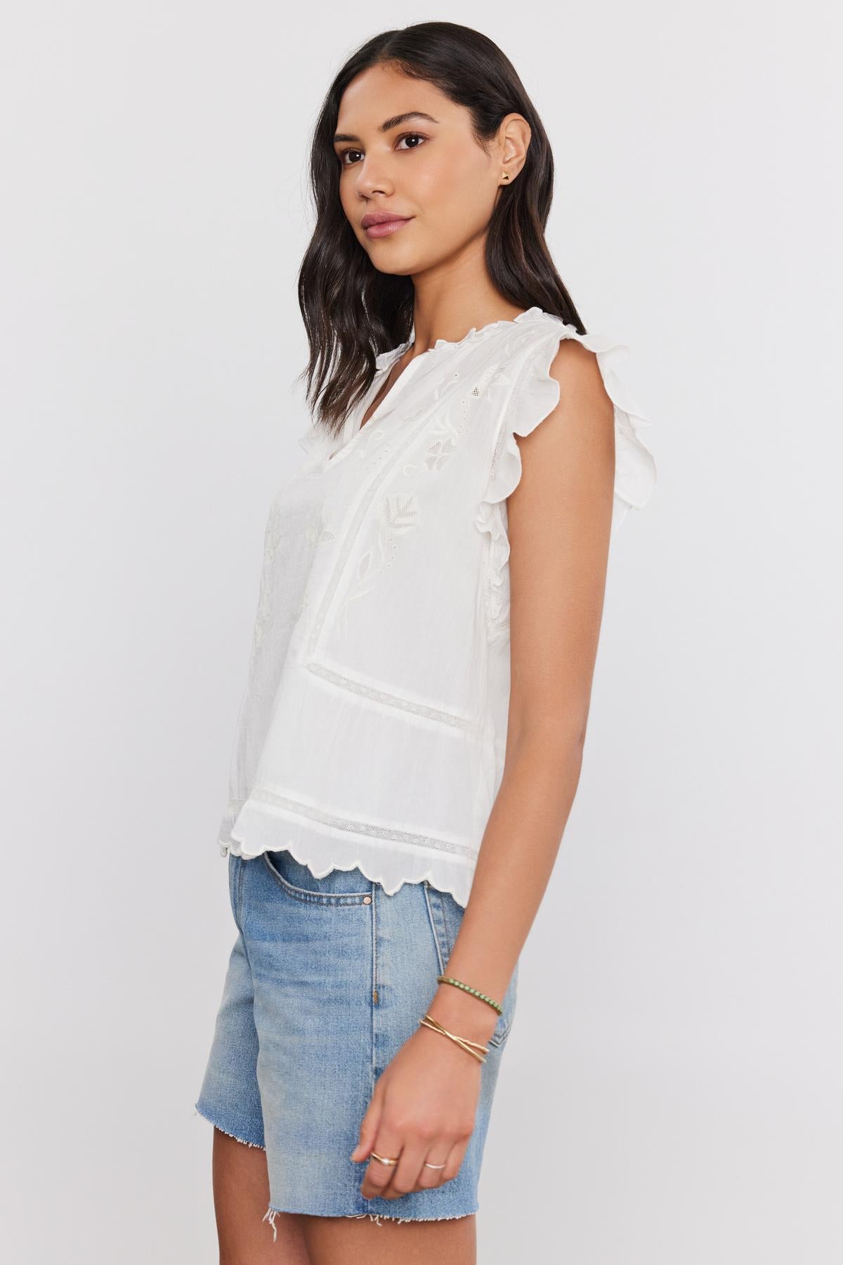 A woman wearing a white CHARLENE TOP with cotton embroidery and denim shorts, standing with a slight smile, facing towards the camera. Brand: Velvet by Graham & Spencer-36830326784193