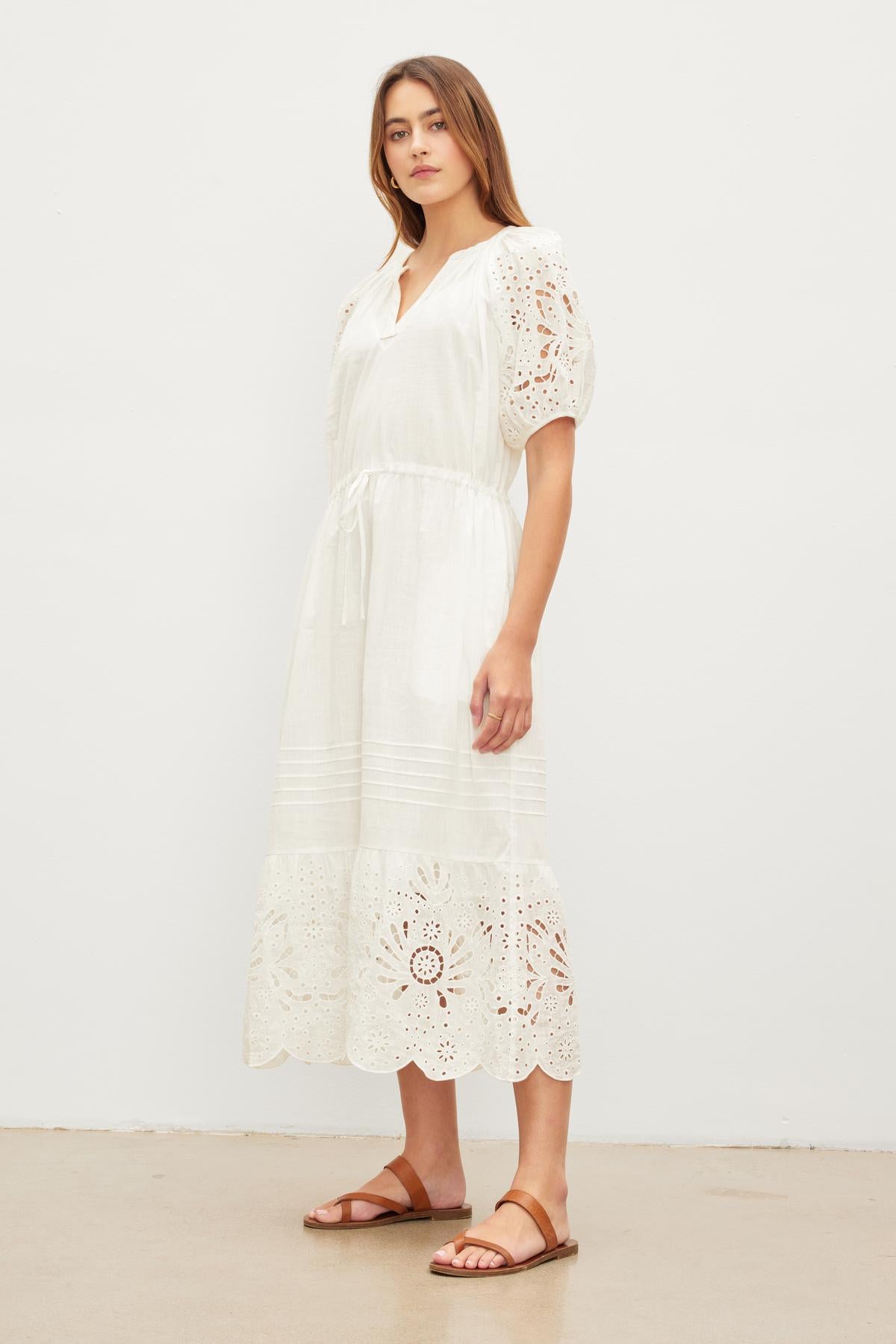   Woman in a white NADIA EMBROIDERED COTTON LACE DRESS with puff sleeves and brown sandals standing against a plain background. (Brand Name: Velvet by Graham & Spencer) 