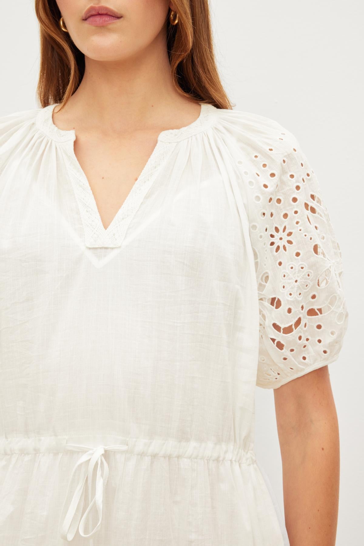   Woman wearing a white NADIA EMBROIDERED COTTON LACE DRESS by Velvet by Graham & Spencer with v-neckline and perforated sleeves, cropped to show from the neck to waist. 