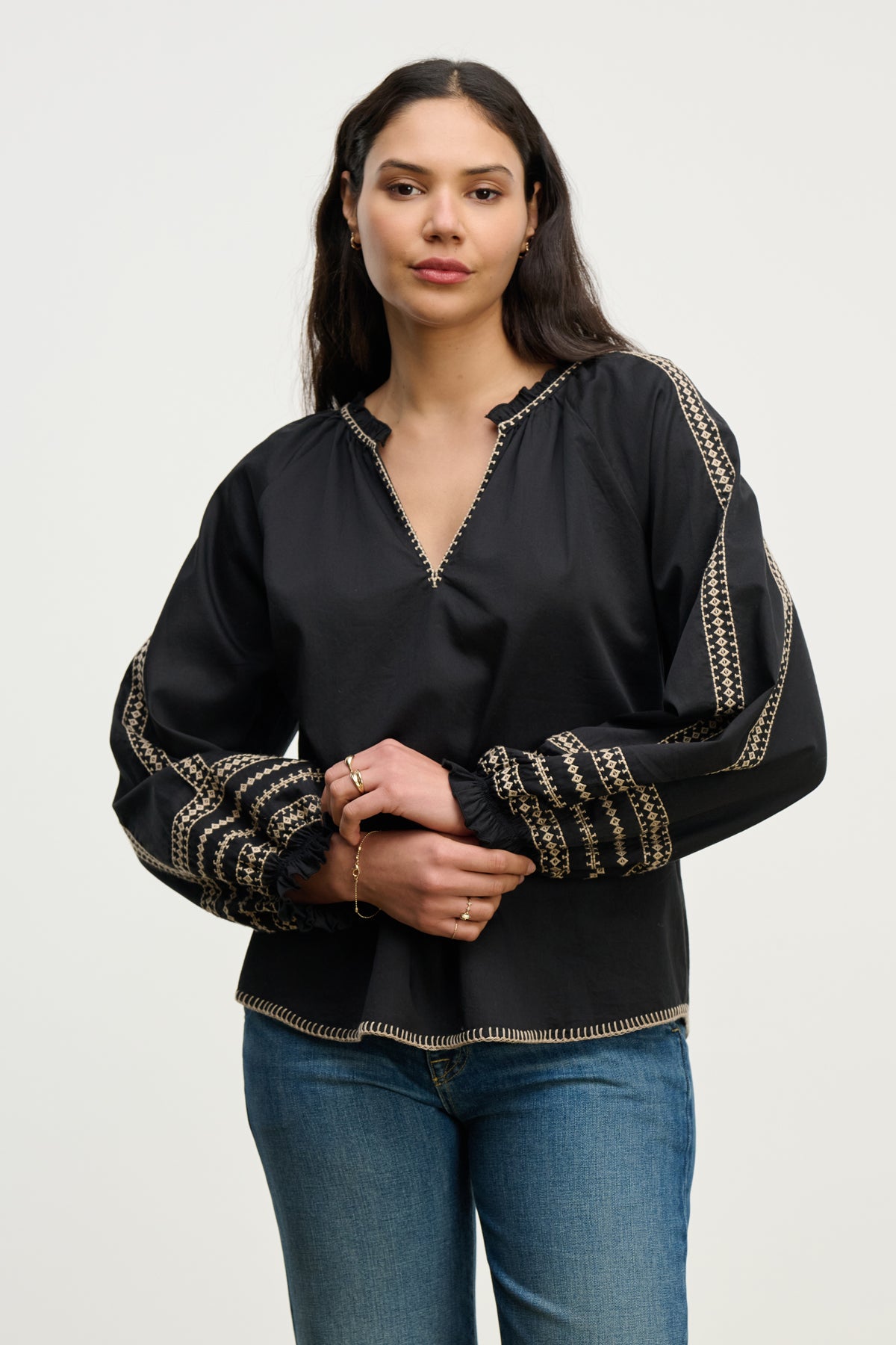 A woman wears the SCARLETT TOP by Velvet by Graham & Spencer, an embroidered blouse with patterned sleeves, paired with blue jeans against a plain light background.