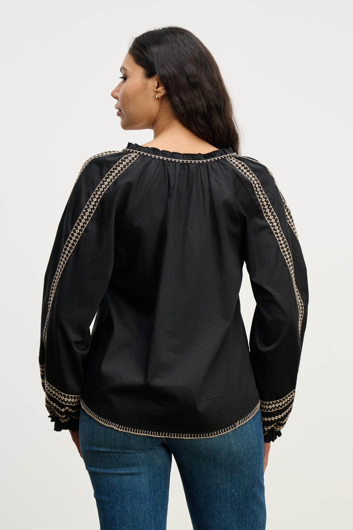 A woman faces away from the camera, wearing the SCARLETT TOP by Velvet by Graham & Spencer—a black embroidered blouse with beige details—paired with blue jeans, set against a plain background.