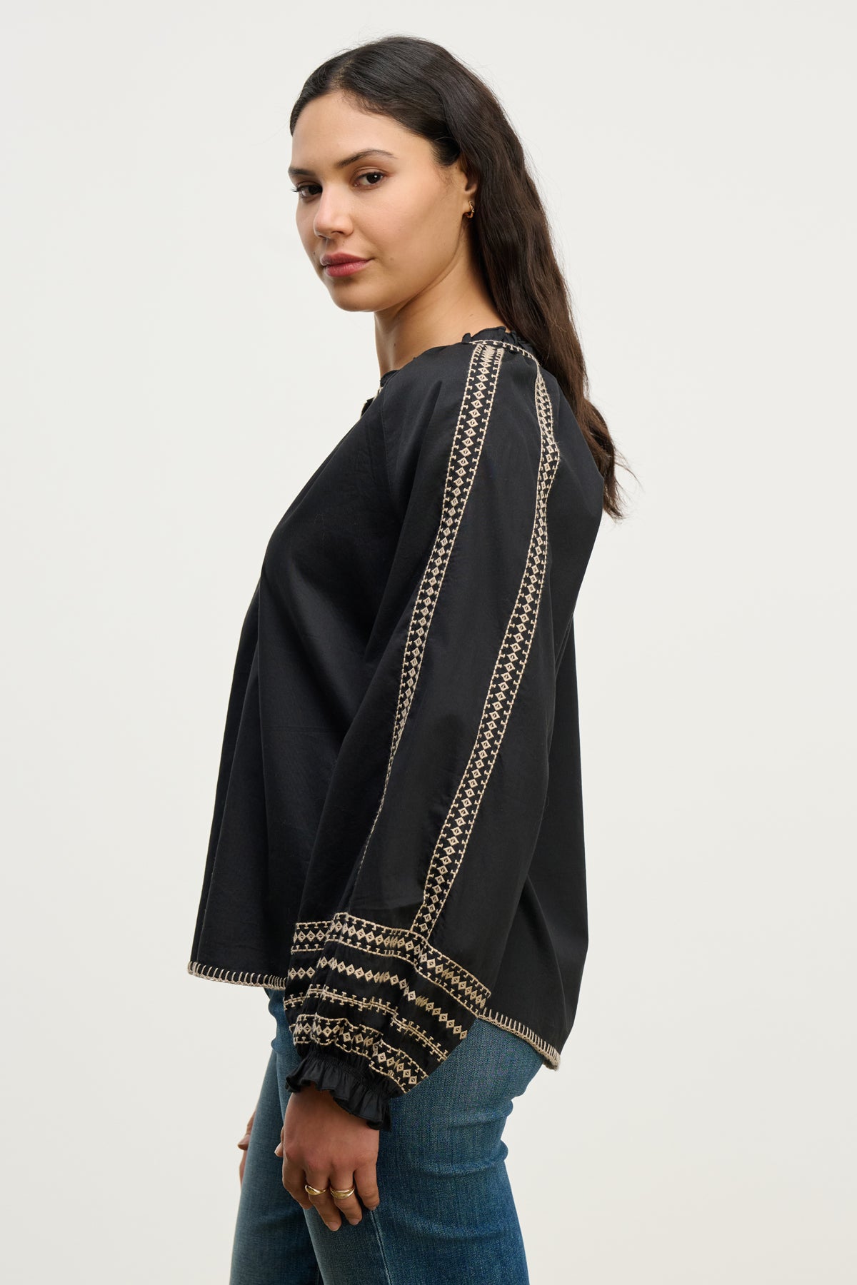 A woman stands in a three-quarter profile wearing the SCARLETT TOP by Velvet by Graham & Spencer, a black embroidered blouse with beige sleeve detailing, paired with blue jeans against a plain light background.