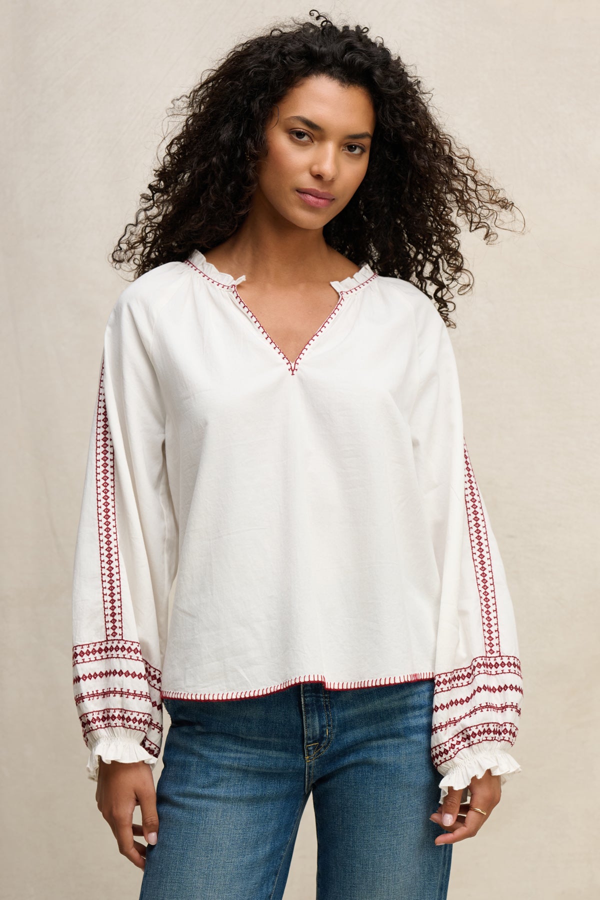 A woman wears the SCARLETT TOP by Velvet by Graham & Spencer, a loose-fitting cotton poplin blouse with red embroidery on the sleeves and cuffs, paired with blue jeans for effortless elegance against a neutral background.-39617777434817