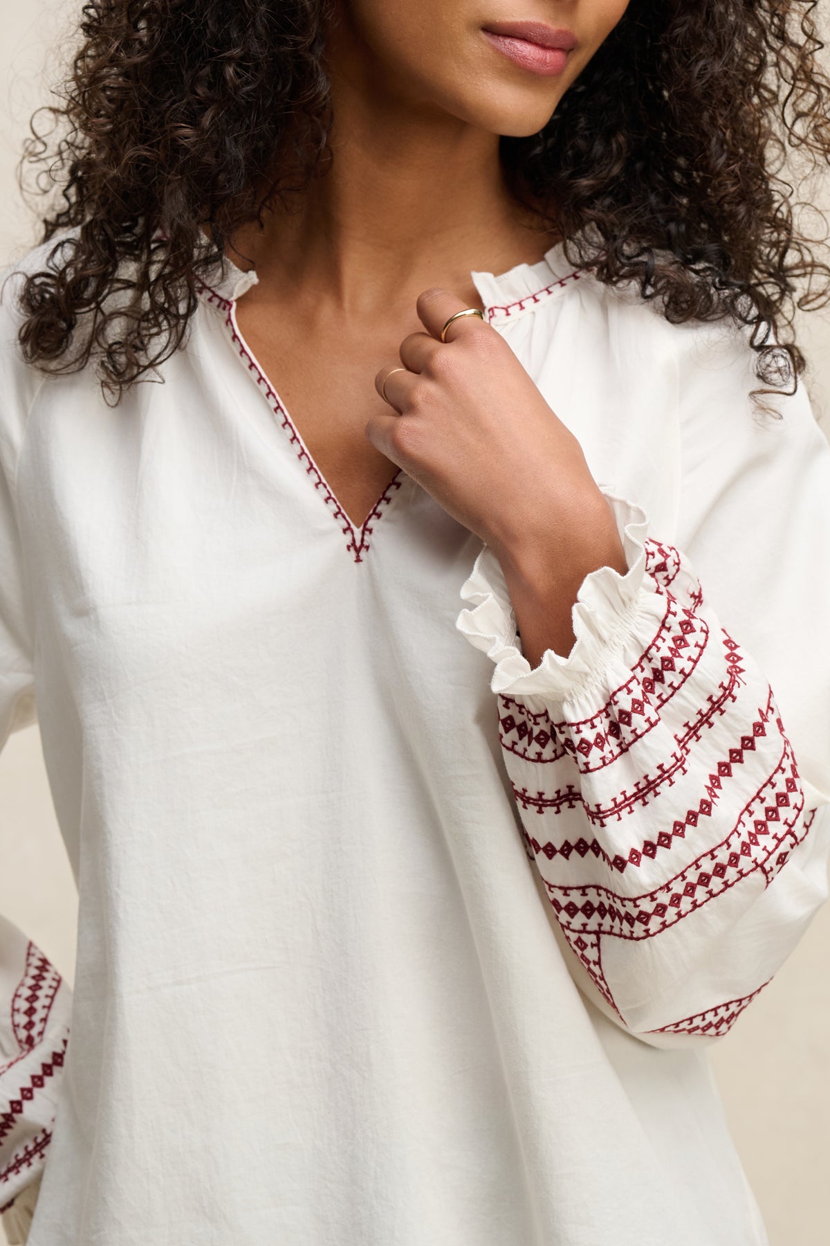 A woman wearing the SCARLETT TOP by Velvet by Graham & Spencer—a white blouse with red embroidery on the sleeves and neckline—holds her hand near her collar.-39617777303745