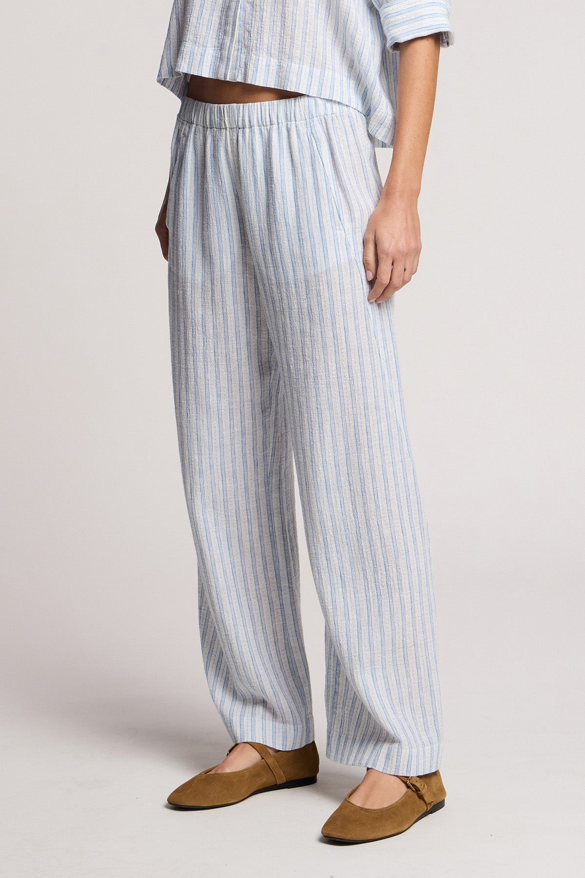 A person wears the EVONY PANT by Velvet by Graham & Spencer—loose-fitting, light blue and white cotton-linen pants with embroidered stripes and a matching top, styled with brown flats against a plain background.-41645018349761