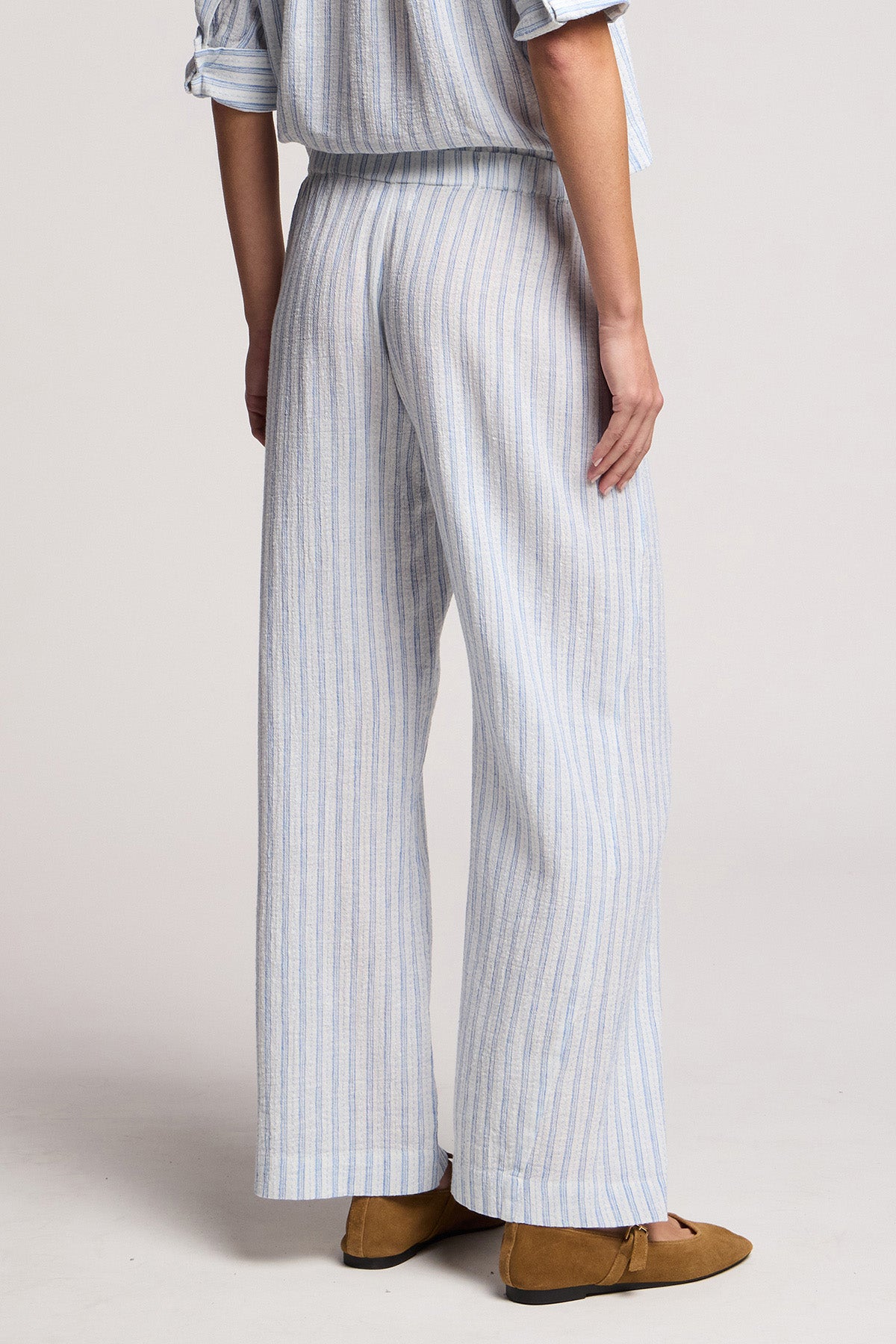 Wearing the EVONY PANT by Velvet by Graham & Spencer in light blue and white cotton-linen with a wide leg silhouette and embroidered stripes, paired with a long-sleeved top and brown flats, shown from the back.-41645018382529