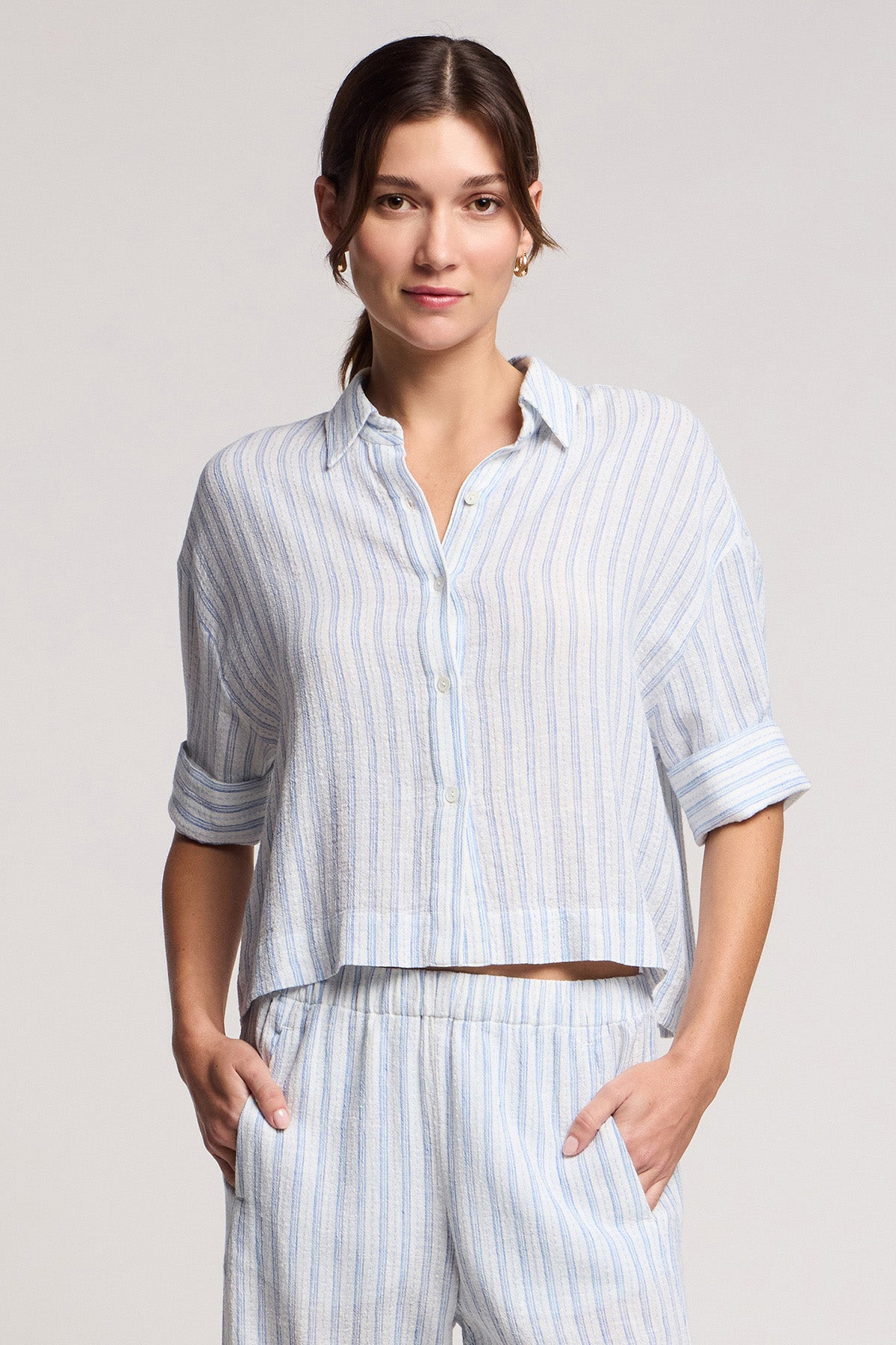 A woman wearing the GREYA SHIRT by Velvet by Graham & Spencer, a light blue and white striped cotton-linen button-up with rolled sleeves, and matching pants, stands with hands in pockets against a plain background.-41645048398017