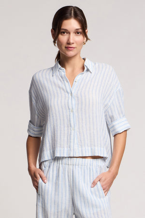 A woman wearing the GREYA SHIRT by Velvet by Graham & Spencer, a light blue and white striped cotton-linen button-up with rolled sleeves, and matching pants, stands with hands in pockets against a plain background.