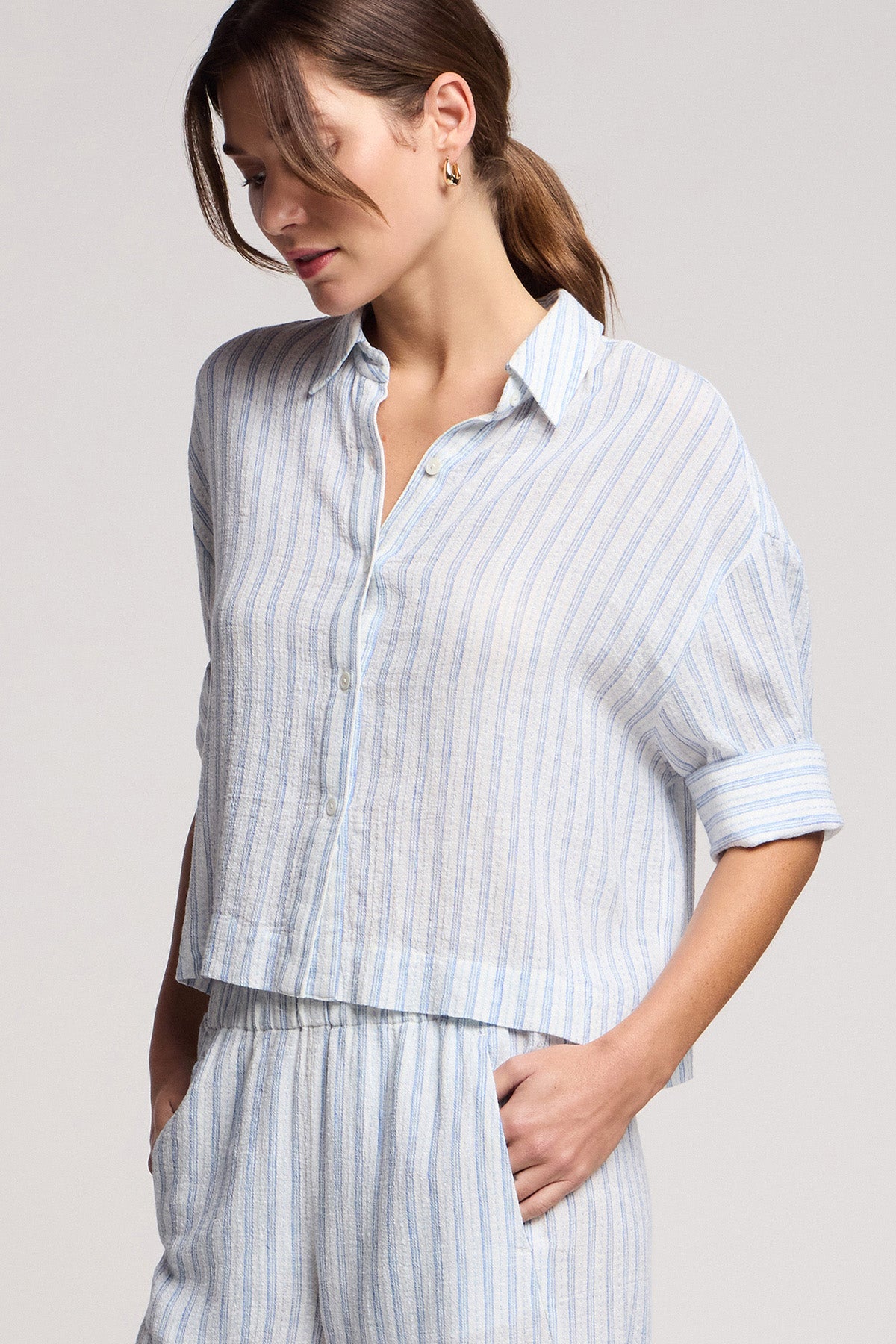 A woman models the GREYA SHIRT by Velvet by Graham & Spencer, a light blue and white striped button-up with rolled sleeves in a cotton-linen blend, paired with matching pants and hands in pockets against a plain background.