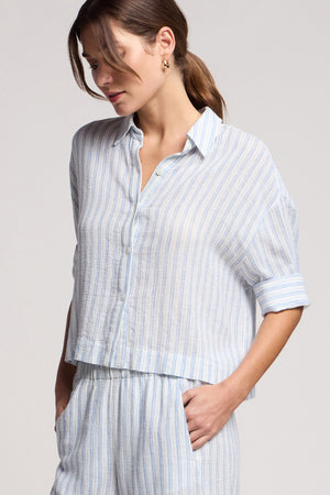 A woman models the GREYA SHIRT by Velvet by Graham & Spencer, a light blue and white striped button-up with rolled sleeves in a cotton-linen blend, paired with matching pants and hands in pockets against a plain background.