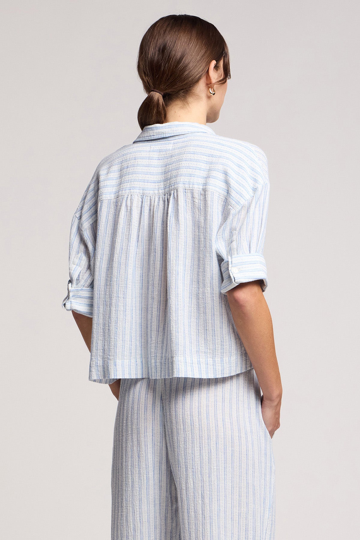 A person shown from the back, hair in a low ponytail, wears the light blue and white striped GREYA SHIRT by Velvet by Graham & Spencer, crafted from a soft cotton-linen blend.-41645048463553